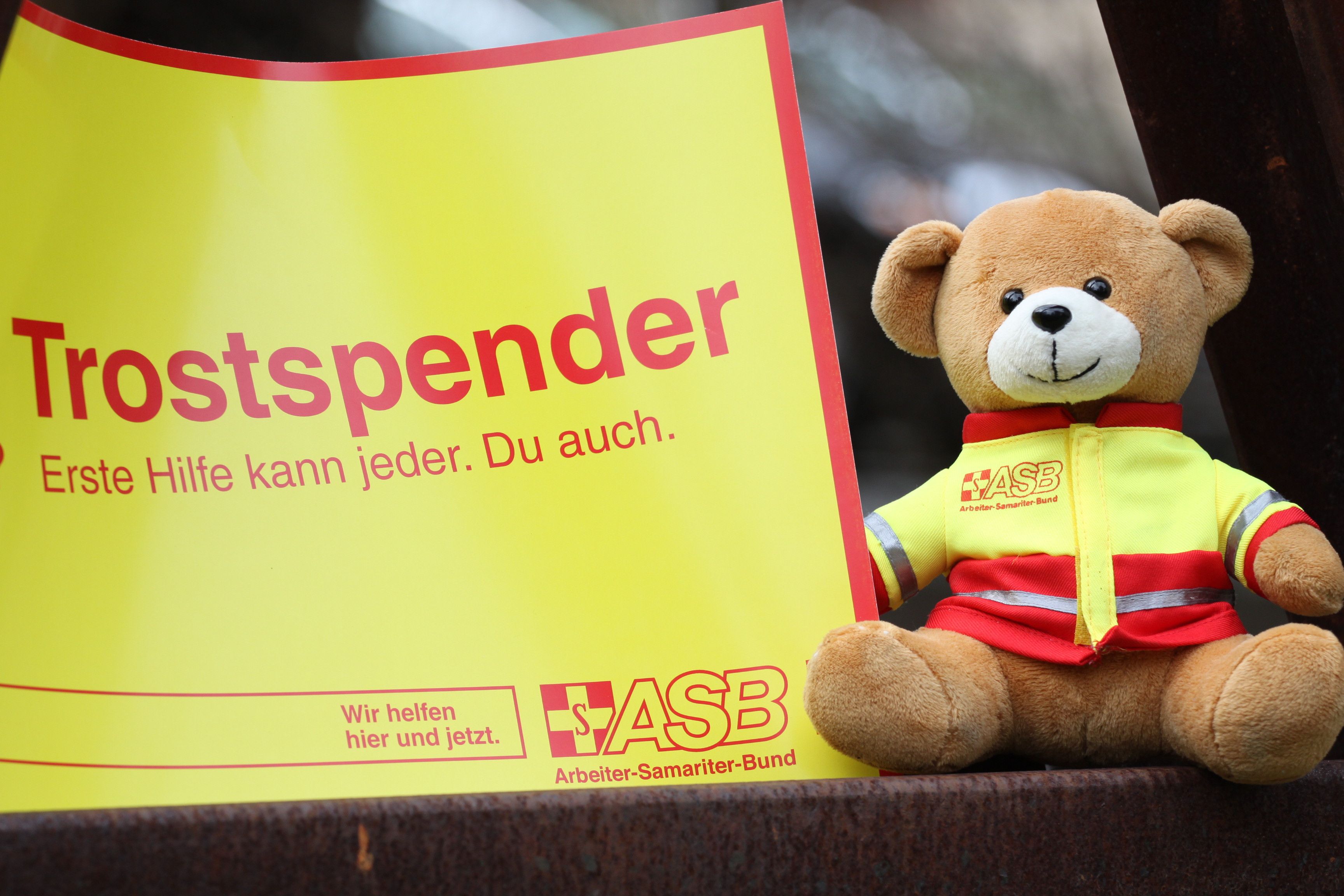 ASB-Teddy neben einem Schild auf dem steht: Trostspender. Erste Hilfe kann jeder. Du auch.